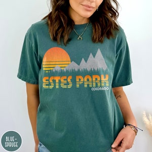 May include: A teal blue t-shirt with a vintage-style graphic of mountains, a sunset, and trees. The text "ESTES PARK COLORADO" is printed on the shirt.