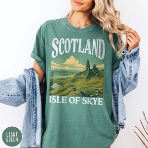 Puede incluir: Camiseta verde claro con la palabra "SCOTLAND" en la parte superior y "ISLE OF SKYE" debajo de una ilustraci&oacute;n de paisaje. La ilustraci&oacute;n representa monta&ntilde;as, un lago y colinas en tonos verdes y marrones.