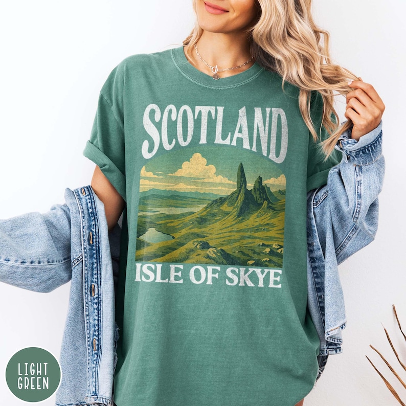 Puede incluir: Camiseta verde claro con la palabra "SCOTLAND" en la parte superior y "ISLE OF SKYE" debajo de una ilustraci&oacute;n de paisaje. La ilustraci&oacute;n representa monta&ntilde;as, un lago y colinas en tonos verdes y marrones.