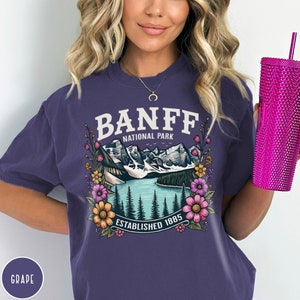 May include: Purple short-sleeved t-shirt with a graphic of a mountain lake scene with the text "Banff National Park Established 1885" surrounded by flowers.