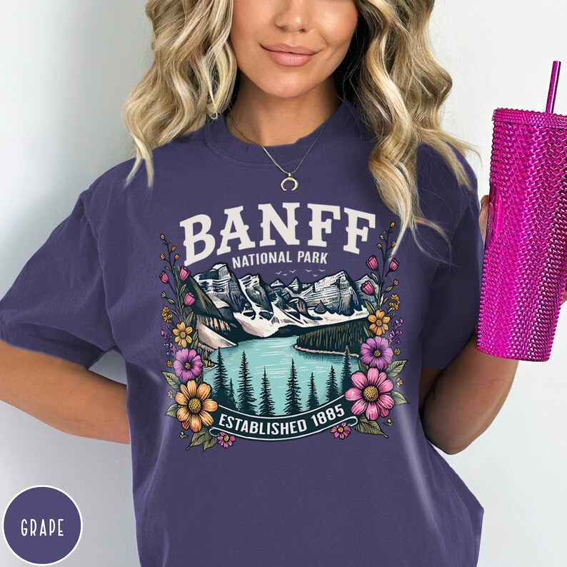 May include: Purple short-sleeved t-shirt with a graphic of a mountain lake scene with the text "Banff National Park Established 1885" surrounded by flowers.