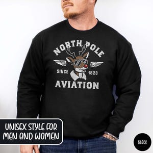 May include: Black crew neck sweatshirt with a distressed graphic featuring a reindeer wearing sunglasses and the text "NORTH POLE AVIATION SINCE 1823." The sweatshirt has the text "UNISEX STYLE FOR MEN AND WOMEN" in a white box.