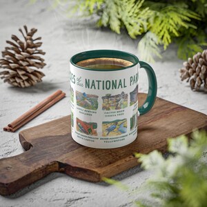May include: A white ceramic mug with a green rim and handle. The mug features a colorful illustration of birds and plants from different national parks in the United States. The text "Birds of our National Parks" is printed on the mug.