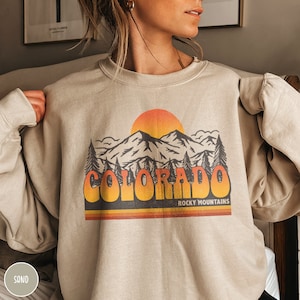 May include: A beige sweatshirt with a vintage-style graphic of mountains, a sunset, and the text "COLORADO ROCKY MOUNTAINS".
