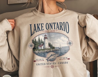 Pode incluir: Uma camisola de gola redonda cor de areia com uma impressão gráfica de um farol numa costa rochosa, um veleiro e um peixe. O texto diz Lake Ontario, Est. 1656, 43°30'N, 77°30'W, United States e Canada, com detalhes de rosas dos ventos.