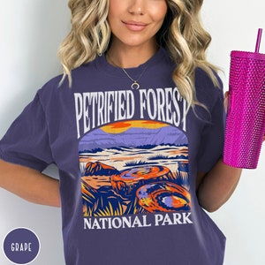 May include: A purple t-shirt with a graphic of a sunset over a landscape with the text "Petrified Forest National Park" in white.