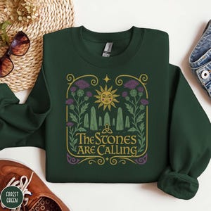May include: Forest green sweatshirt with a graphic design featuring a smiling sun, standing stones, and purple thistles. The text "The Stones Are Calling" is in a decorative font. The sweatshirt is made of a soft material.