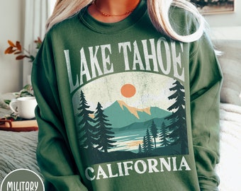 May include: A green sweatshirt with a vintage-style graphic of Lake Tahoe, California. The graphic features a mountain range, a lake, trees, and a setting sun.