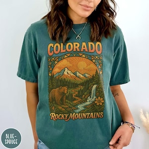 May include: Teal-colored t-shirt featuring a vintage-style graphic of the Colorado Rocky Mountains. The design includes a bear, mountains, a waterfall, and the words "COLORADO" and "Rocky Mountains" in orange and white.