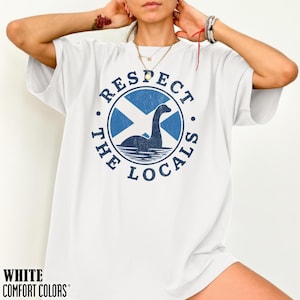 May include: White t-shirt with a blue graphic featuring a stylized Loch Ness Monster inside a circle with a Scottish flag design. The text "RESPECT THE LOCALS" is printed around the circle. The words "WHITE COMFORT COLORS" are printed at the bottom.