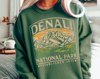 May include: A green sweatshirt with a vintage-style graphic of Denali National Park. The graphic features a mountain range with a sunset in the background and the text "Denali National Park Established 1917".