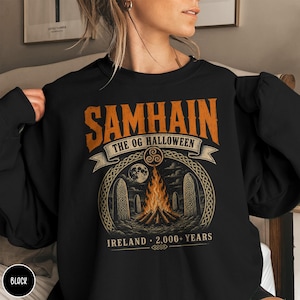 May include: A black crewneck sweatshirt featuring a graphic print with the orange text SAMHAIN THE OG HALLOWEEN. The design includes a central bonfire, standing stones, a full moon, and the text IRELAND 2,000+ YEARS in a circular frame.