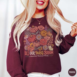 May include: Maroon sweatshirt with a floral design featuring various flowers and butterflies. The text "Let Our Parks Bloom" and "America's National Parks" are printed below the floral design. The sweatshirt is a solid maroon color.