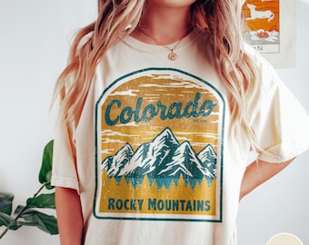 May include: A white t-shirt with a vintage-style graphic of the Rocky Mountains and the text "Colorado est. 1876" and "Rocky Mountains". The shirt is a light ivory color.