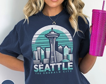 May include: A navy blue t-shirt with a vintage-style graphic of the Seattle skyline, including the Space Needle. The text "Seattle The Emerald City" is printed below the graphic.