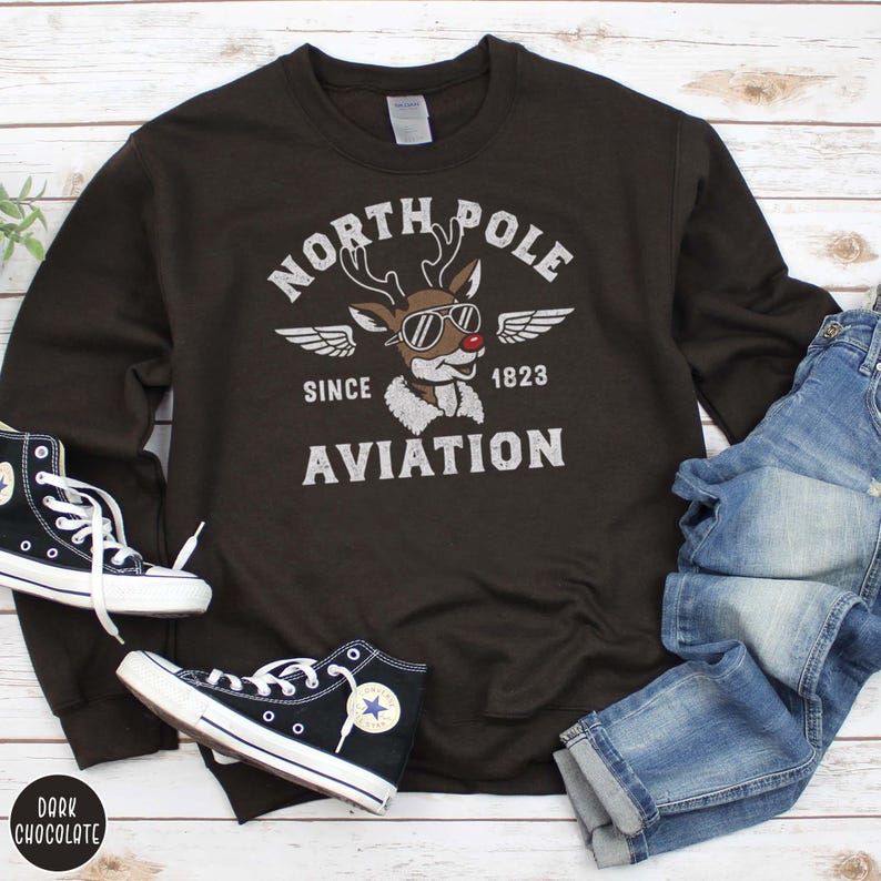 May include: Dark chocolate-colored sweatshirt with a graphic featuring a reindeer wearing sunglasses and the text "North Pole Aviation Since 1823." The image includes black high-top sneakers and blue jeans.
