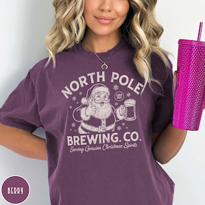 May include: Berry-colored t-shirt with a graphic of Santa Claus giving a thumbs up and holding a beer, with the text "NORTH POLE BREWING CO." and "Serving Genuine Christmas Spirits."