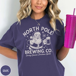 May include: A purple t-shirt with a graphic of Santa Claus giving a thumbs up and holding a beer. The text on the shirt reads "NORTH POLE BREWING CO. Serving Genuine Christmas Spirits" in white. A pink tumbler is held in the right hand.