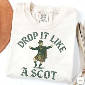Puede incluir: Camiseta de color marfil con el texto "DROP IT LIKE A SCOT" en verde. La camiseta presenta un gr&aacute;fico de un hombre con vestimenta tradicional escocesa. La camiseta est&aacute; doblada y tiene una etiqueta que dice "Comfort Colors".