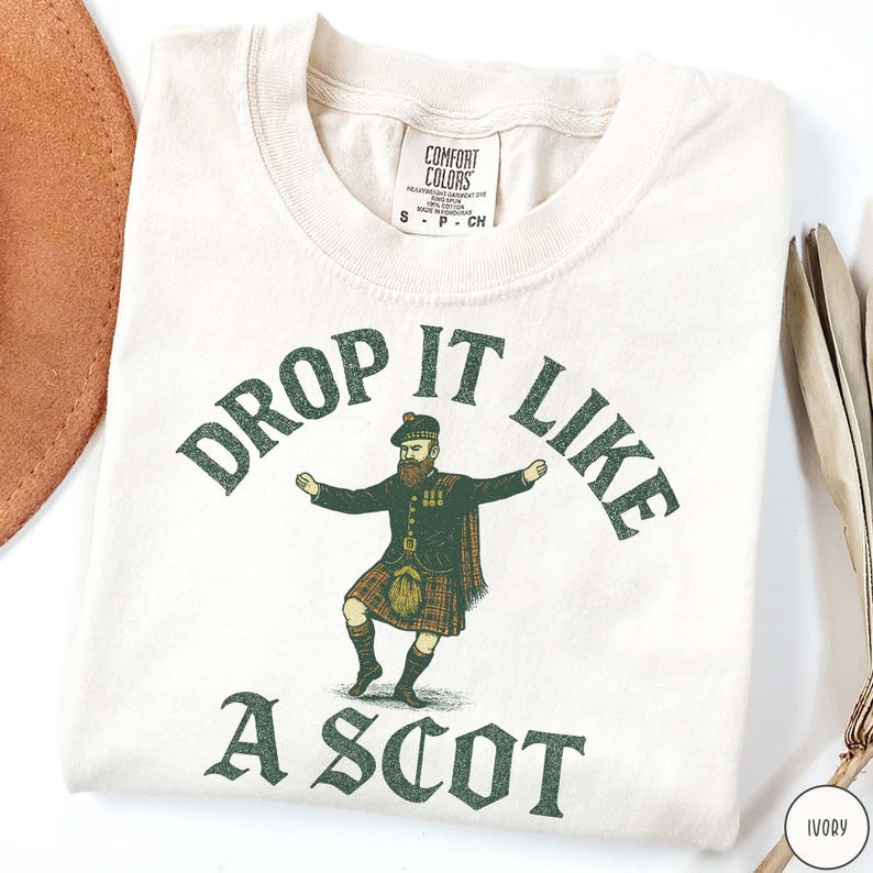Puede incluir: Camiseta de color marfil con el texto "DROP IT LIKE A SCOT" en verde. La camiseta presenta un gr&aacute;fico de un hombre con vestimenta tradicional escocesa. La camiseta est&aacute; doblada y tiene una etiqueta que dice "Comfort Colors".
