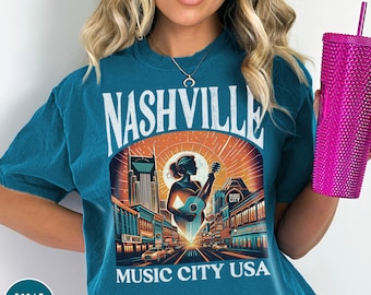 May include: A teal blue t-shirt with a vintage-style graphic of the Nashville skyline. The graphic features a woman playing a guitar in front of the city's iconic buildings. The text "Nashville Music City USA" is printed below the graphic.