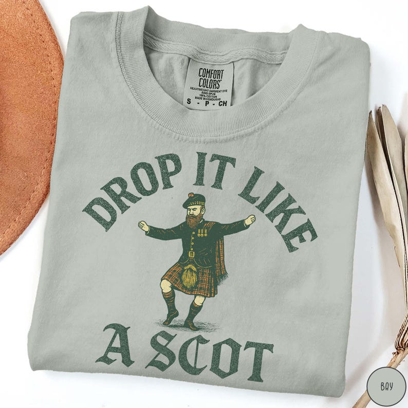 Puede incluir: Camiseta gris claro con la frase "DROP IT LIKE A SCOT" en verde. La camiseta presenta una ilustraci&oacute;n de un hombre con kilt. La camiseta est&aacute; hecha de un material suave y tiene cuello redondo.
