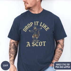 Puede incluir: Camiseta azul marino con la frase "DROP IT LIKE A SCOT" y una ilustraci&oacute;n de un hombre con kilt. El dise&ntilde;o tiene un aspecto vintage. La camiseta est&aacute; etiquetada como "DENIM" y "UNISEX STYLE FOR MEN AND WOMEN".