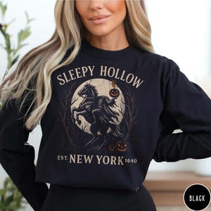 May include: Black long-sleeve shirt featuring a graphic of the Headless Horseman from Sleepy Hollow. The design includes a horse, a pumpkin, and the text "Sleepy Hollow Est. New York 1640". The shirt is a solid black color.