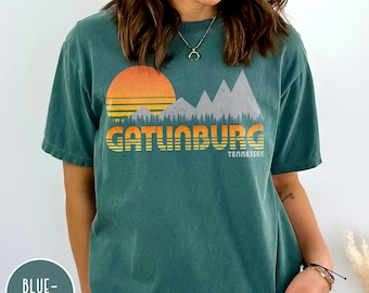 May include: A teal blue t-shirt with a vintage-style graphic of mountains, a sunset, and the text "Gatlinburg Tennessee".