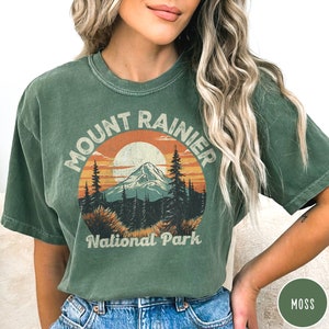 May include: A green t-shirt with a vintage-style graphic of Mount Rainier National Park. The graphic features a mountain range, a sunset, and the words "Mount Rainier National Park".