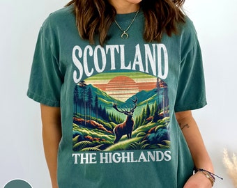 May include: A green t-shirt with a vintage-style graphic of a deer standing in a forest with mountains and a sunset in the background. The text "Scotland The Highlands" is printed below the graphic.