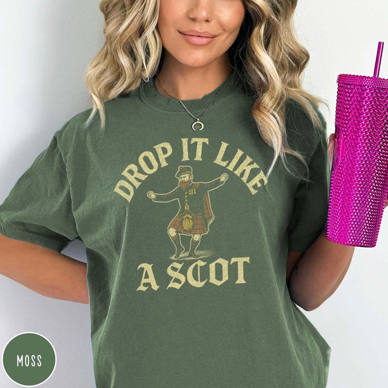 Puede incluir: Camiseta verde musgo con el texto "DROP IT LIKE A SCOT" y un gr&aacute;fico de un hombre bailando con un kilt. La camiseta tiene mangas cortas y cuello redondo. Una taza rosa se sostiene en la mano derecha.