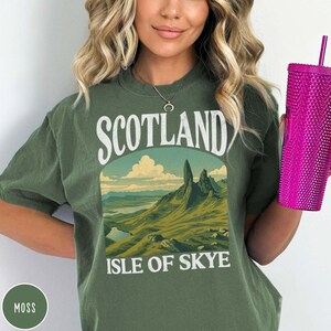 Puede incluir: Camiseta verde musgo con un gr&aacute;fico de paisaje de la Isla de Skye, Escocia. El gr&aacute;fico presenta monta&ntilde;as, un lago y el texto "SCOTLAND ISLE OF SKYE" en blanco. En la imagen se sostiene un vaso rosa texturizado.