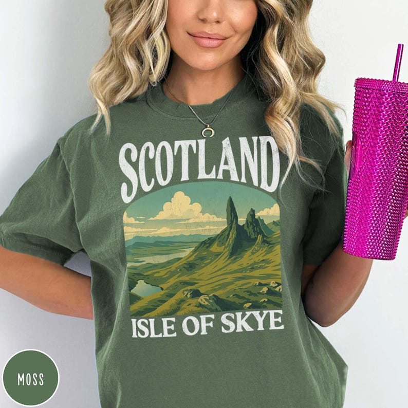 Puede incluir: Camiseta verde musgo con un gr&aacute;fico de paisaje de la Isla de Skye, Escocia. El gr&aacute;fico presenta monta&ntilde;as, un lago y el texto "SCOTLAND ISLE OF SKYE" en blanco. En la imagen se sostiene un vaso rosa texturizado.
