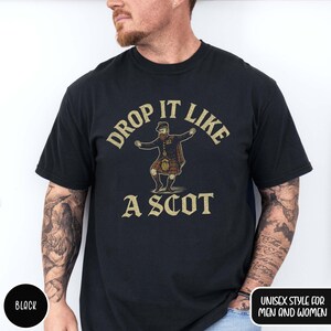 Puede incluir: Camiseta negra con texto dorado "DROP IT LIKE A SCOT". El dise&ntilde;o muestra un personaje de dibujos animados con kilt bailando. La camiseta est&aacute; etiquetada como "UNISEX STYLE FOR MEN AND WOMEN".