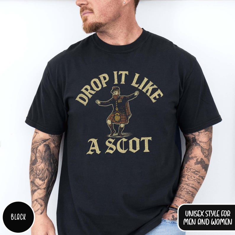 Puede incluir: Camiseta negra con texto dorado "DROP IT LIKE A SCOT". El dise&ntilde;o muestra un personaje de dibujos animados con kilt bailando. La camiseta est&aacute; etiquetada como "UNISEX STYLE FOR MEN AND WOMEN".