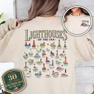 May include: Beige sweatshirt featuring a hand-drawn illustration of 30 lighthouses of the USA. The design includes the text "LIGHTHOUSES OF THE USA" and a small graphic of a lighthouse. A tag reads "INCLUDES 30 HAND-DRAWN LIGHTHOUSES OF THE USA."