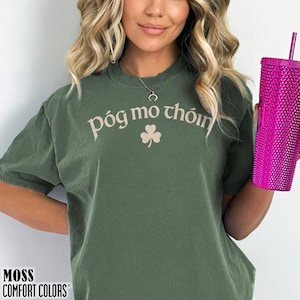 May include: A moss green short-sleeve t-shirt featuring the text póg mo thóin in a light beige font arched above a small shamrock graphic. The shirt is paired with a bright pink textured tumbler that has a matching straw extending from the top.