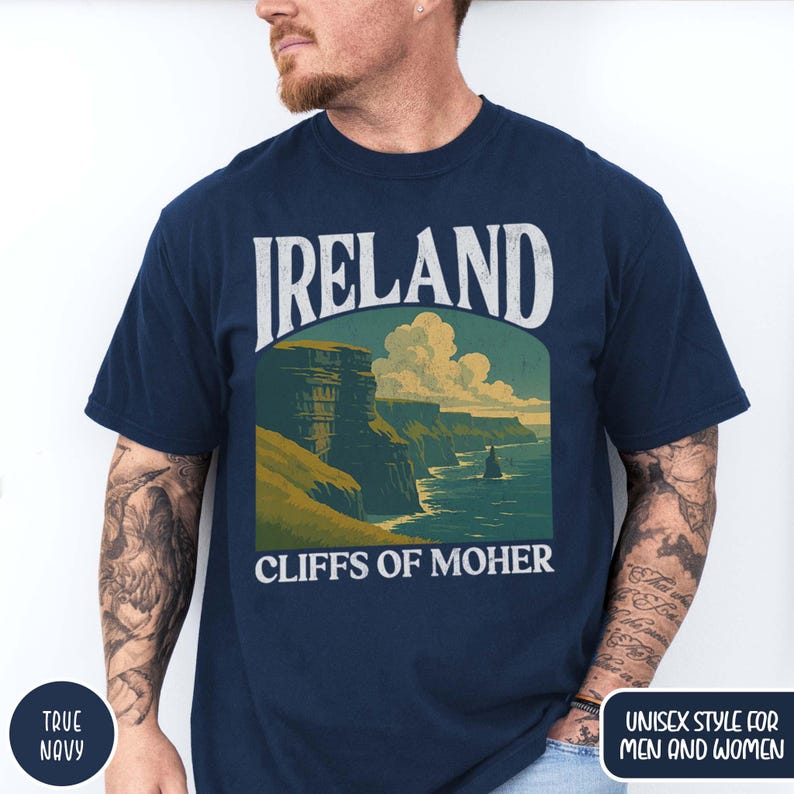 May include: A navy blue t-shirt with the word "IRELAND" in white block letters above a scenic illustration of the Cliffs of Moher. The words "CLIFFS OF MOHER" are below the illustration. The shirt has a round neck and short sleeves.