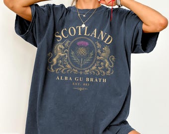 Puede incluir: Una camiseta azul marino de manga corta con un gráfico dorado de un unicornio y un león flanqueando un cardo morado. El texto dice SCOTLAND, ALBA GU BRÀTH y EST. 843. El diseño está centrado en el pecho de la prenda.