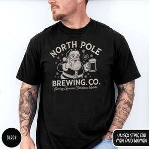 May include: Black t-shirt with a graphic featuring Santa Claus giving a thumbs up and holding a beer mug. The text reads "NORTH POLE BREWING CO. Serving Genuine Christmas Spirits" in a vintage style. The shirt is labeled "UNISEX STYLE FOR MEN AND WOMEN."
