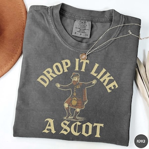 Puede incluir: Camiseta gris oscuro con la frase dorada "DROP IT LIKE A SCOT". El dise&ntilde;o incluye un hombre con vestimenta escocesa tradicional. Una cadena dorada con un colgante est&aacute; sobre la camiseta. La camiseta tiene una etiqueta Comfort Colors.