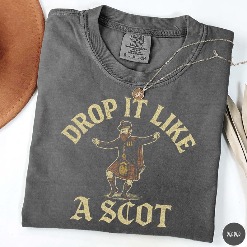 Puede incluir: Camiseta gris oscuro con la frase dorada "DROP IT LIKE A SCOT". El dise&ntilde;o incluye un hombre con vestimenta escocesa tradicional. Una cadena dorada con un colgante est&aacute; sobre la camiseta. La camiseta tiene una etiqueta Comfort Colors.