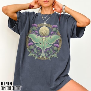 May include: A charcoal gray short-sleeve t-shirt featuring a central graphic of a pale green luna moth centered within a circular gold Celtic knot border, surrounded by purple thistle flowers and a full moon against a dark background.