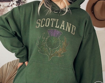 Pode incluir: Moletom verde militar com a palavra "SCOTLAND" em letras douradas desgastadas acima de um gráfico de cardo. O cardo tem detalhes roxos e verdes. O moletom tem capuz com cordão e um bolso frontal.