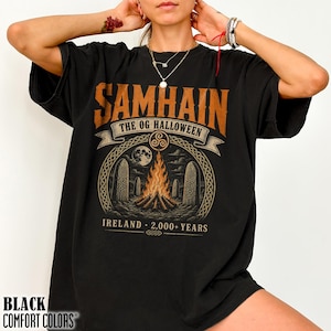 May include: A black t-shirt featuring a graphic print with the text SAMHAIN THE OG HALLOWEEN IRELAND 2,000+ YEARS. The design includes a central bonfire, standing stones, a full moon, and a triskelion symbol, all rendered in orange and cream tones.