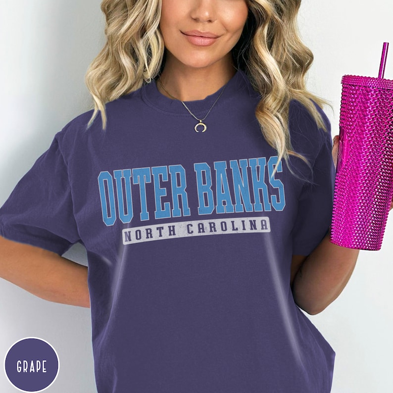 Op de afbeelding: Een paars T-shirt met de tekst "Outer Banks North Carolina" in blauwe en witte letters.