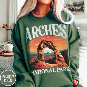 May include: A green sweatshirt with a vintage-style graphic of Arches National Park. The graphic features a sunset scene with a large rock arch and the text "ARCHES NATIONAL PARK".
