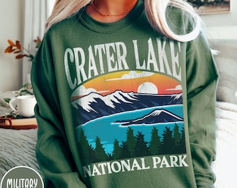 May include: A green sweatshirt with a vintage-style graphic of Crater Lake National Park. The graphic features a sunset over a mountain range with a lake in the foreground. The text "Crater Lake National Park" is printed in white below the graphic.