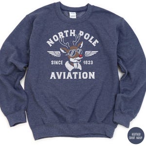 May include: A navy blue sweatshirt with a graphic featuring a reindeer wearing sunglasses and the words "NORTH POLE AVIATION SINCE 1823." The design includes wings and a vintage aesthetic. The sweatshirt is a heather sport navy color.